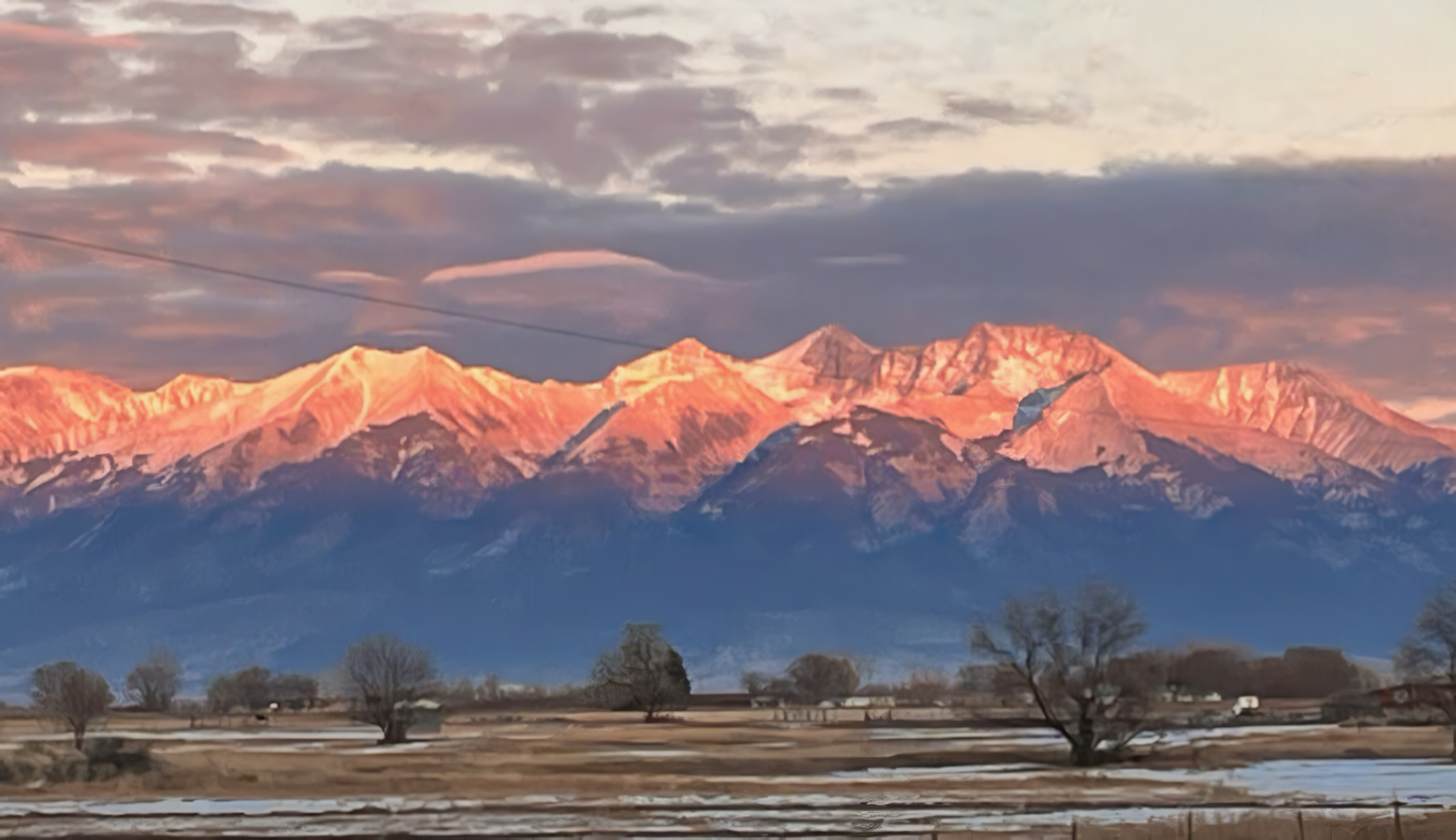 Snow-capped mountains at sunset with a foreground of bare trees and open fields, under a partly cloudy sky.